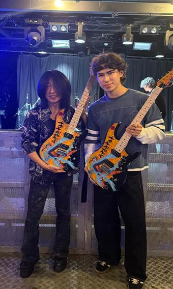 Dos personas en un escenario, cada una con una guitarra Naruto Shippuden, identificadas como Yasuharu Takanashi y Billy Smith de Vader Guitars.