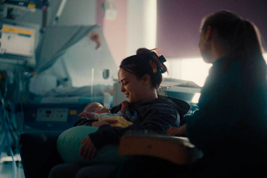 Mujer joven sonriendo a un bebé prematuro en brazos con sonda nasal, junto a monitores y una persona de espaldas