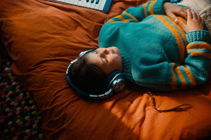 Niña tumbada en una cama con funda naranja, auriculares puestos y ojos cerrados junto a un teclado azul