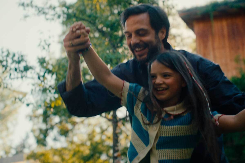 Un hombre con barba y una niña ríen y bailan cogidos de la mano en un jardín con árboles iluminados por luz otoñal.
