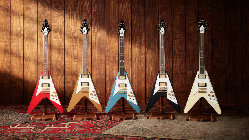 Cinco guitarras Flying V en pie sobre soportes de madera, con acabados rojo, dorado, azul claro, negro y blanco.