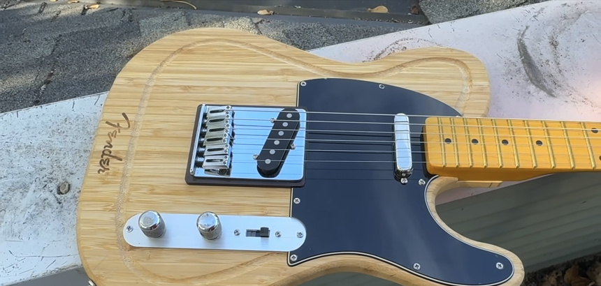 Guitarra terminada con cuerpo de tabla Fender Telecaster, puente cromado, pastillas, golpeador negro y placa de controles sobre fondo al aire libre.